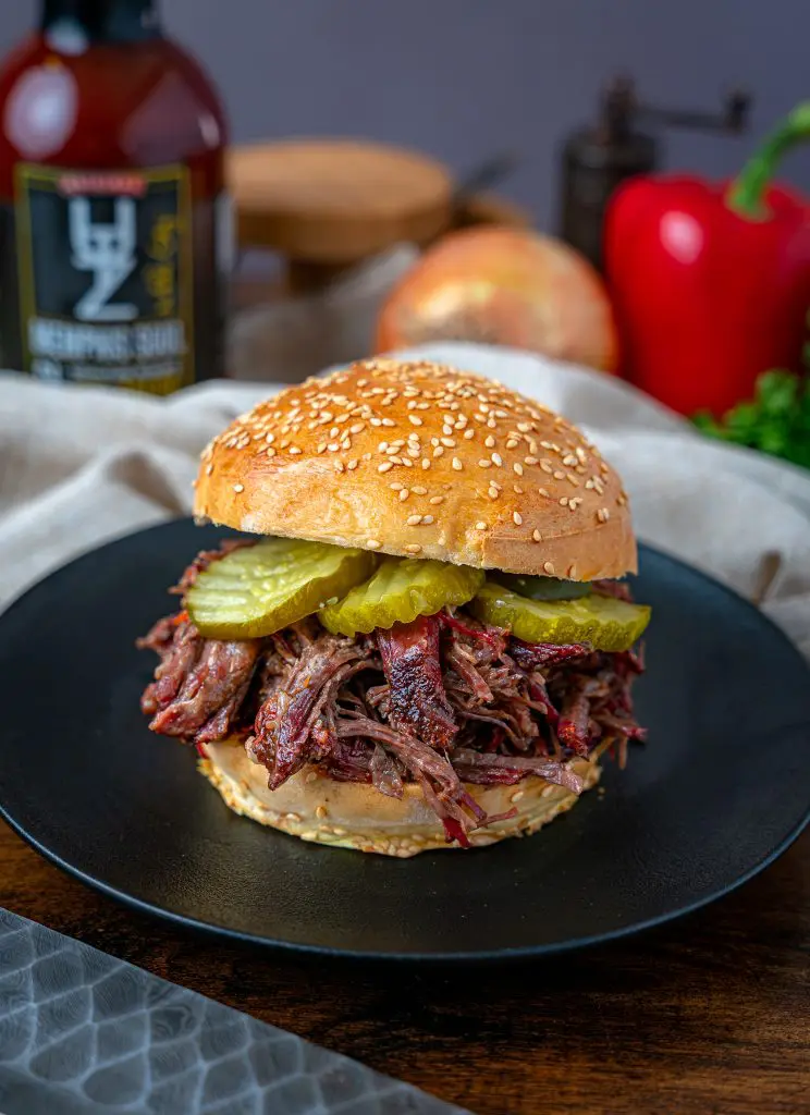 Shredded chuck roast sandwich with bbq sauce, onions and peppers in the background.