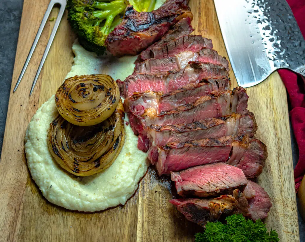 sliced ribeye on a cutting board with grilled onions and mashed potatoes
